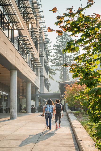 Image shows two students walking at the south-western corner of the Behavioral Social Science building. It is a smoky fall day. 